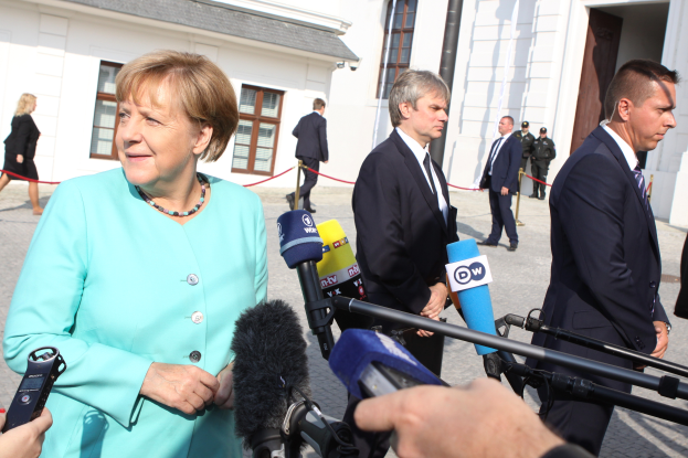 Bundeskanzlerin Angela Merkel spricht vor der Bundeskanzlei in Berlin mit Reportern, umgeben von Menschen mit Mikrofonen und Gebäuden im Hintergrund.