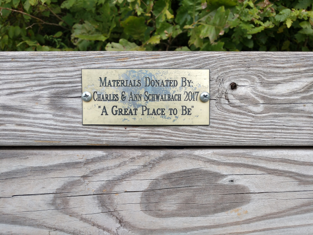 Eine hölzerne Bank mit einer Plakette, auf der "Spende von Charles & Ann Schwabach 2017: Ein schöner Ort zu sein" steht, vor üppigen grünen Pflanzen.