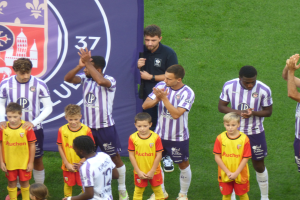 Eine Gruppe junger Jungs auf einem Fußballfeld stehend, die Hände klatschen, mit einem Banner und Logos im Hintergrund und saftigem grünem Gras darunter.