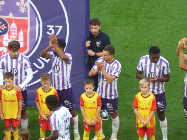 Eine Gruppe junger Jungs auf einem Fußballfeld stehend, die Hände klatschen, mit einem Banner und Logos im Hintergrund und saftigem grünem Gras darunter.