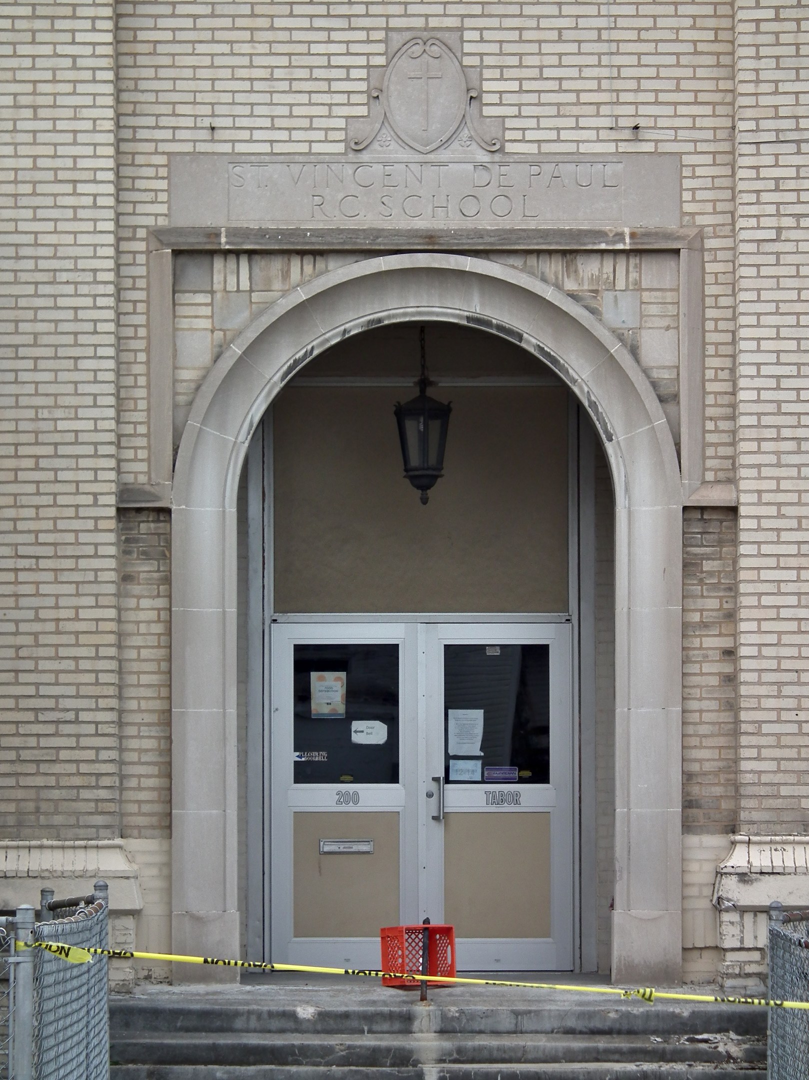 Ein Eingang eines Backsteingebäudes mit gelbem Absperrband, Treppenstufen mit Maschendrahtzaun auf beiden Seiten, eine Tür mit angeklebten Papieren, eine hängende Lampe und eine Wandinschrift, die wahrscheinlich den Namen der Schule angibt.