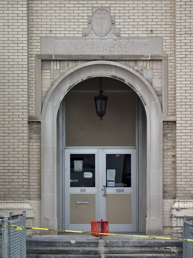 Ein Eingang eines Backsteingebäudes mit gelbem Absperrband, Treppenstufen mit Maschendrahtzaun auf beiden Seiten, eine Tür mit angeklebten Papieren, eine hängende Lampe und eine Wandinschrift, die wahrscheinlich den Namen der Schule angibt.