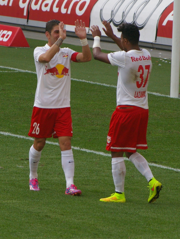 Zwei Fußballspieler stehen auf einem grünen Feld und klatschen sich ab, mit einem Pfahl und einer Tafel mit Text im Hintergrund.
