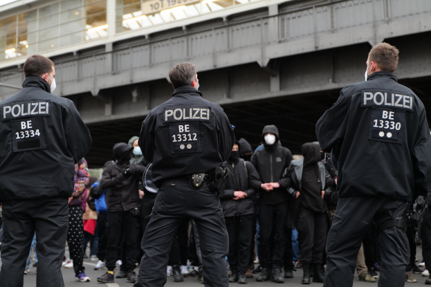 Eine Gruppe von Polizisten in Uniform steht vor einer Menge von Menschen in schwarzen Uniformen und Masken, mit einer Brücke und einem Gebäude im Hintergrund, während einer Protestaktion in einer Stadt.