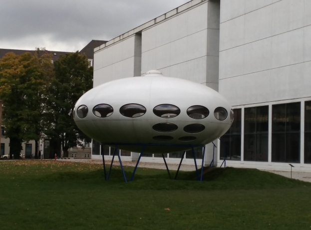 Eine große weiße UFO-Skulptur thront auf einem grünen Feld, umgeben von Gebäuden, Bäumen und Fahrzeugen unter einem bewölkten Himmel.
