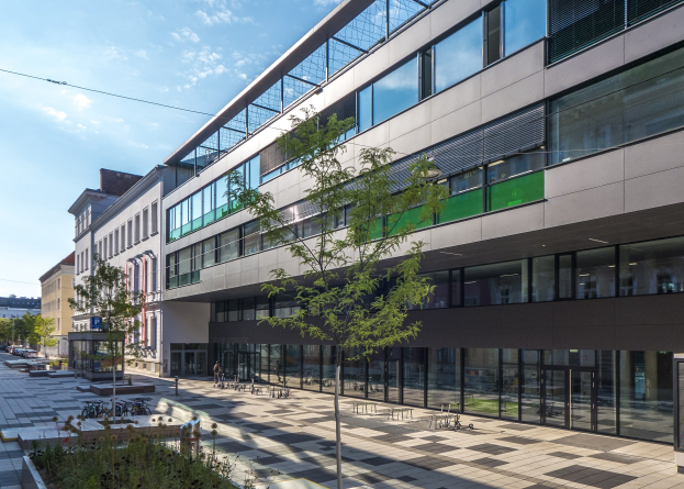 Großes modernes Gebäude mit zahlreichen Fenstern, umgeben von Bäumen und Grünflächen, mit Menschen und Fahrzeugen im Vordergrund, unter einem bewölkten Himmel.