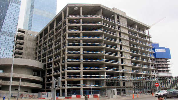 Ein großes Bauwerk im Bau in einer städtischen Umgebung, umgeben von Wolkenkratzern, Fahrzeugen, Fußgängern, Verkehrsinfrastruktur und Baumaschinen gegen einen klaren Himmel.