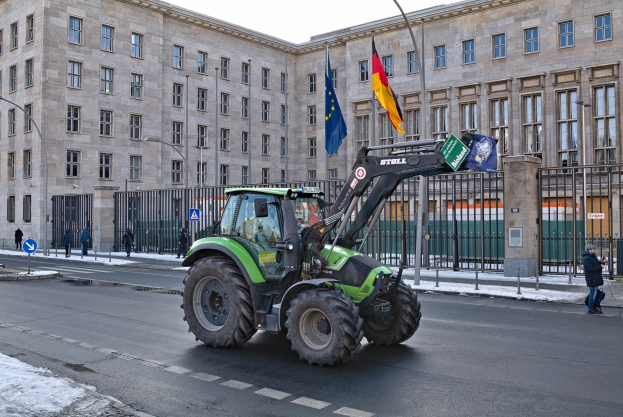 Ein Traktor fährt auf einer Straße vor einem Gebäude, mit einer Person darin. Rechts geht eine Person auf dem Gehweg, links sind Schilder, Pfosten, Laternen und ein Zaun zu sehen. Im Hintergrund sind Gebäude, Flaggen und der Himmel zu erkennen.