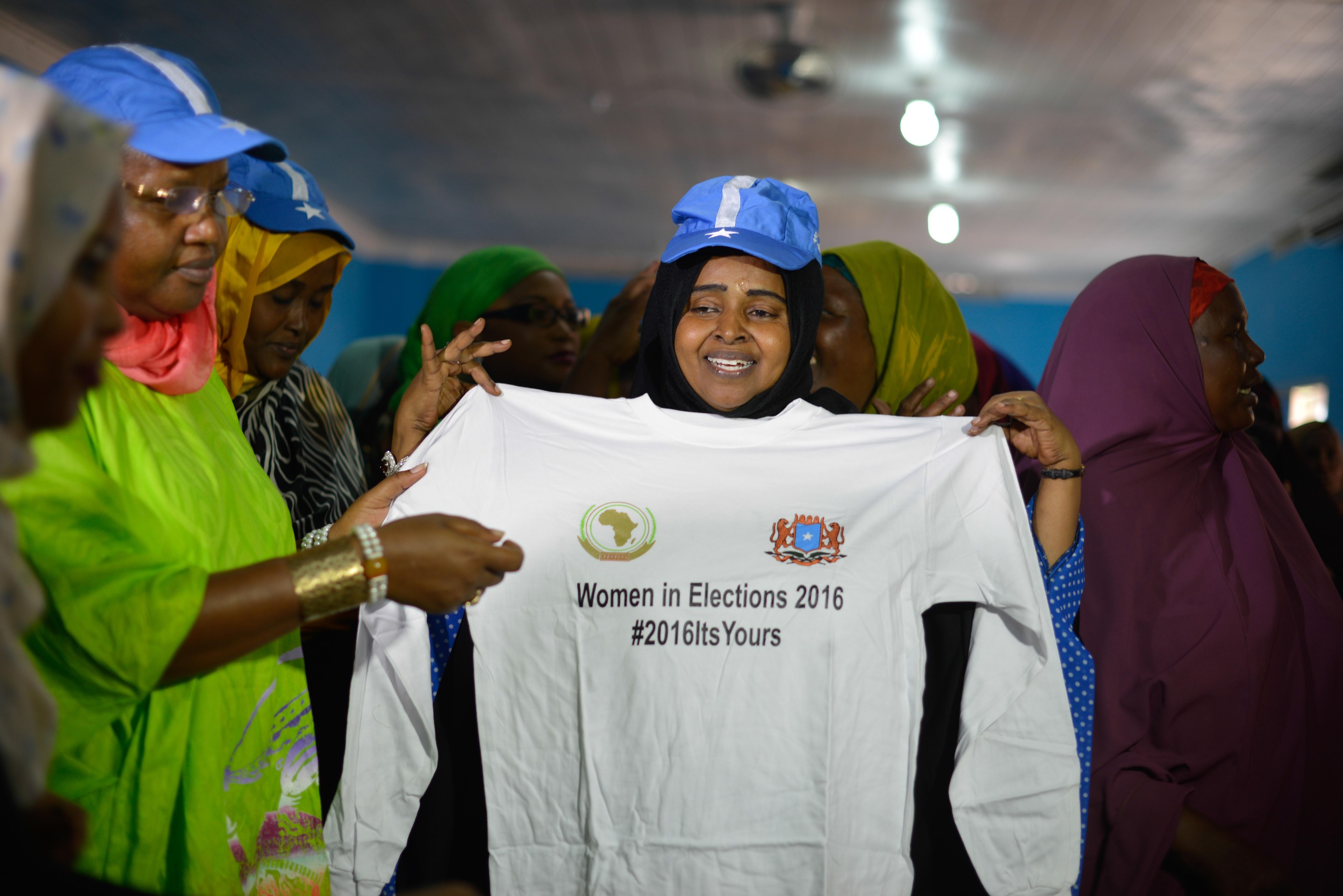 Eine Gruppe von Frauen hält ein weißes T-Shirt mit der Aufschrift "Women in Elections 2016" hoch, einige tragen blaue Mützen, unter Deckenbeleuchtung.