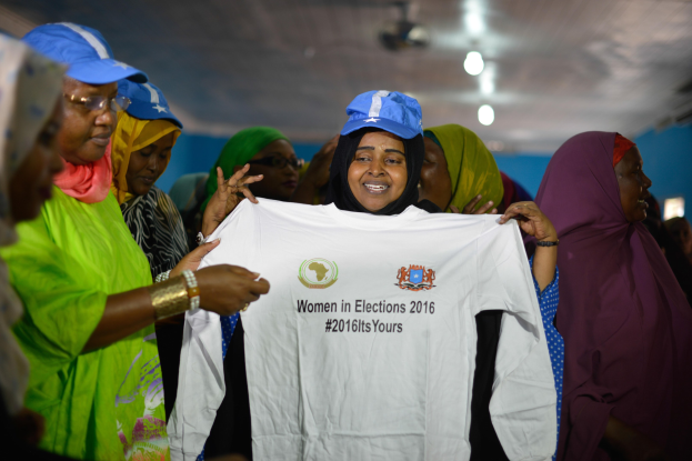 Eine Gruppe von Frauen hält ein weißes T-Shirt mit der Aufschrift "Women in Elections 2016" hoch, einige tragen blaue Mützen, unter Deckenbeleuchtung.