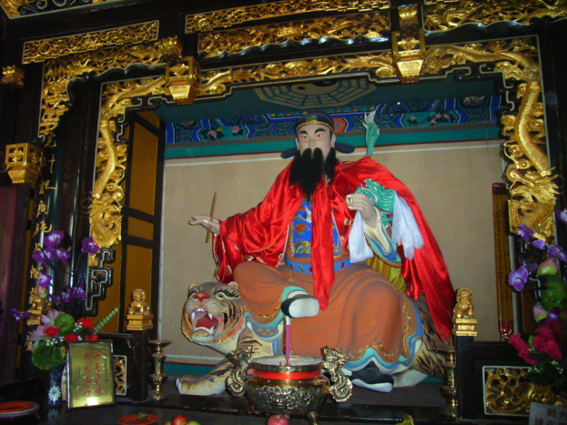 Eine Statue eines chinesischen Mönchs, der mit buntem Stoff geschmückt ist, sitzt auf einer Tigerstatue innerhalb eines Bogens mit goldenen Verzierungen, vor dem eine Vase und in der Mitte eine Schüssel stehen.