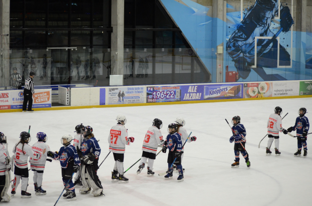 Gruppe junger Menschen, die Eis-hockey auf einer Indoor-Eisfläche spielen, mit Helmen und Hockey-Schlägern, sowie einer Person auf der linken Seite und Bannern, Säulen und Glaswänden im Hintergrund.
