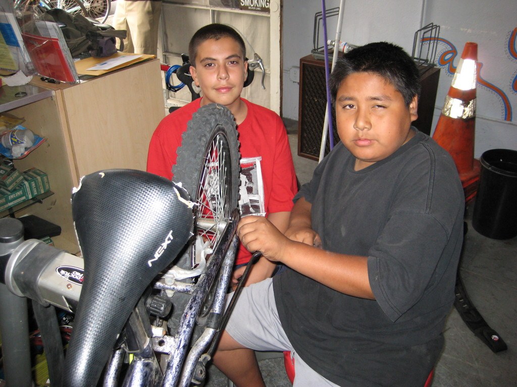 Zwei Personen reparieren ein Fahrrad in der Mitte, mit einem Schreibtisch und verschiedenen Gegenständen links und Fahrrädern und anderen Gegenständen im Hintergrund.