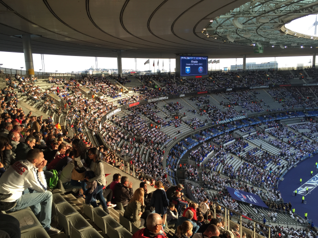 Eine große Menge Menschen in einem Stadion bei einem Fussballspiel, mit einer Bühne auf der rechten Seite, Fahnen, Stangen und einem Bildschirm im Hintergrund, unter einem sichtbaren Himmel, in der Allianz Arena in München, Deutschland.
