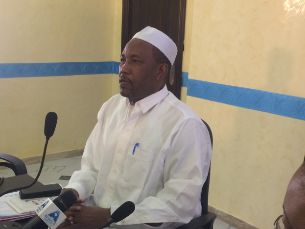 Ein Mann in weißem Hemd und Hut sitzt an einem Tisch mit Mikrofonen, spricht, während Papiere und Gegenstände auf dem Tisch zu sehen sind, und ein weiterer Person mit Brille rechts von ihm in einem Raum mit einer Tür im Hintergrund.