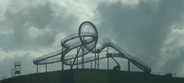 Eine Achterbahn auf dem Gipfel eines Hügels mit einem bewölkten Himmel im Hintergrund, umgeben von Stützpfählen und -strukturen.