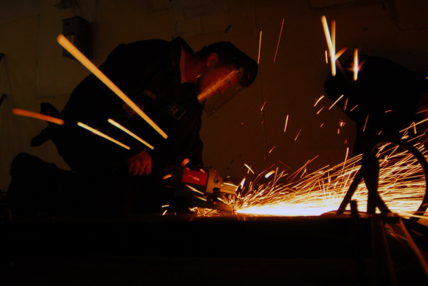 Zwei Männer schweißen ein Metallstück in einem dunklen Raum, Funken fliegen von der Schweißmaschine, mit einer Wand im Hintergrund.