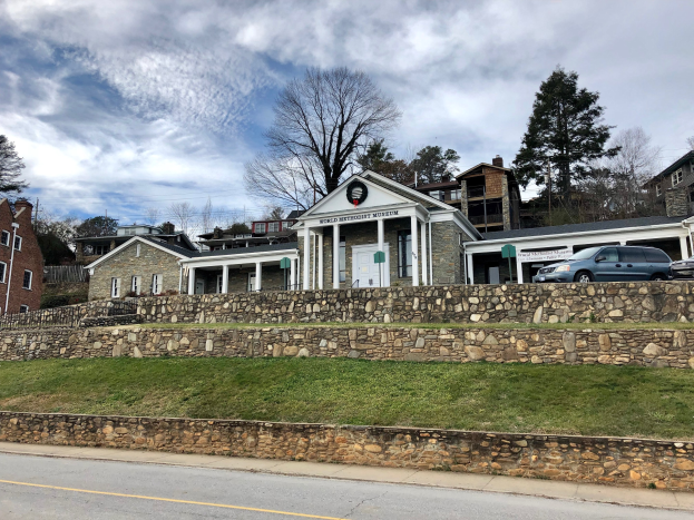 Steinmauer mit parkenden Autos, angrenzende Gebäude mit Fenstern und Türen, Bäume und ein bewölkter Himmel, beschriftet als kürzlich wiedereröffnetes Weltkulturerbe-Museum.