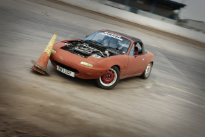 Eine Person fährt ein rotes Auto auf einer Rennstrecke und überholt einen Verkehrskegel mit einem unscharfen Hintergrund, der hohe Geschwindigkeit andeutet.