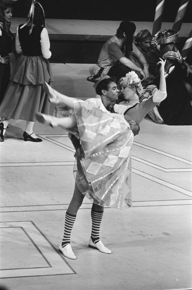 Ein Schwarz-Weiß-Foto eines Paares, das auf einer Bühne tanzt, mit einigen Menschen auf einer Treppe im Hintergrund und einem Pfahl im Vordergrund.