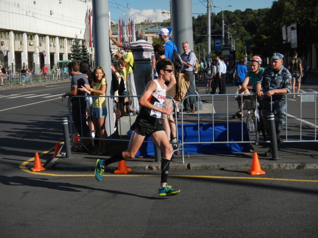 Ein Mann läuft einen Marathon auf einer Stadtstraße, umgeben von Zuschauern auf dem Gehweg, einige halten Fahrräder, mit städtischer Infrastruktur und bewölktem Himmel im Hintergrund.
