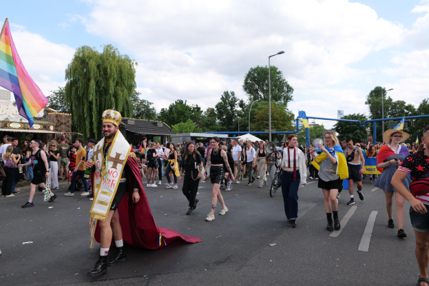 Eine Gruppe von Menschen marschiert in einem Umzug, trägt eine Regenbogenfahne und Musikinstrumente, mit Laternenmasten, Bäumen und Hütten im Hintergrund.