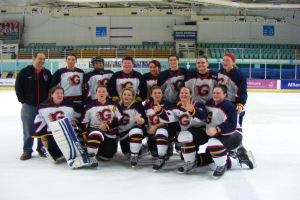 Eine Gruppe von Frauen-Eishockeyspielerinnen in Helmen und Schlittschuhen posiert für ein Foto auf dem Eis, eine hält einen Hockeystock, mit Stühlen, Tafeln mit Text, Geländern und Deckenlampen im Hintergrund.
