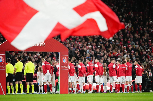 Gruppe von Menschen auf einem Fussballfeld mit einer roten und weißen Flagge im Vordergrund, einem Bogen mit der Aufschrift "Bayern München vs Bayern München Wetten & Vorschau" im Hintergrund und einer großen Menge im Stadion.
