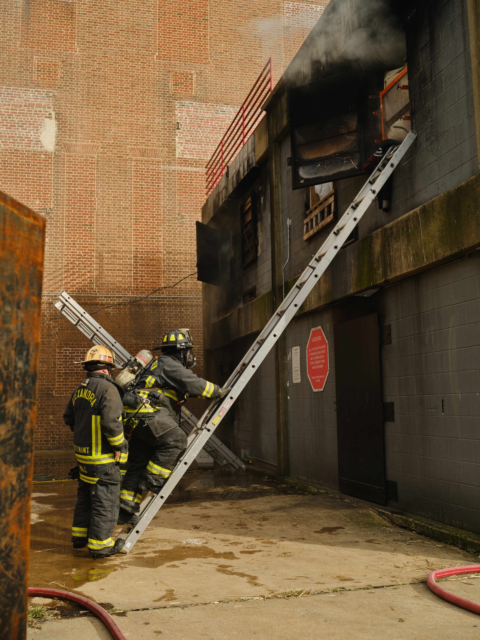 Feuerwehrleute mit Helmen klettern an einer Leiter zu einem raucherfüllten Gebäude hoch, während ein metallisches Objekt und Bodenschläuche zu sehen sind.