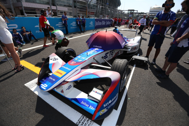 Formula E-Rennwagen mit weißer, blauer, roter und schwarzer Lackierung, der auf einer Strecke geparkt ist, umgeben von Menschen, mit einem Schirm im Vordergrund und Rennstrecken-Zaun, Gebäuden, Bäumen und einem klaren Himmel im Hintergrund.