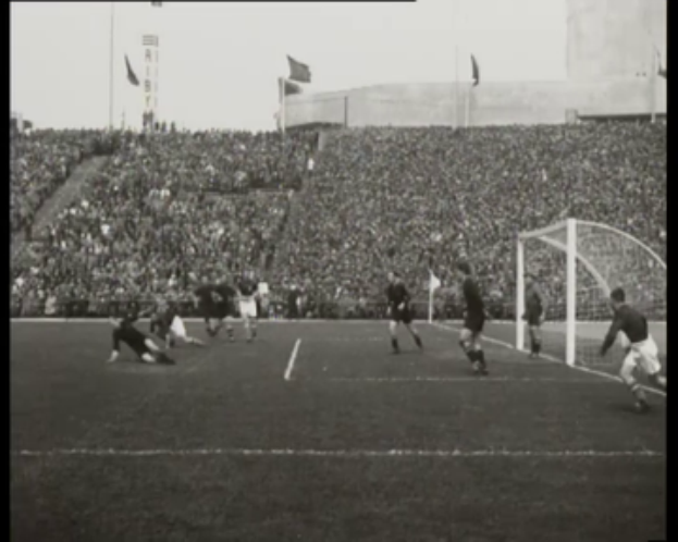 Ein Schwarz-Weiß-Foto eines Fußballspiels der WM 1958 zwischen Manchester United und Liverpool, mit Spielern auf dem Feld, einem Torpfosten auf der rechten Seite, Zuschauern in den Rängen und Fahnen im Hintergrund.