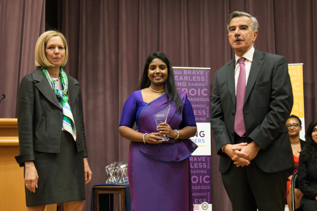 Zwei Frauen und ein Mann stehen auf einer Bühne mit einem braunen Vorhang dahinter, die Frau in der Mitte hält einen Award, mit mehreren Awards auf einem Tisch davor und einem Podium mit einem Mikrofon links.