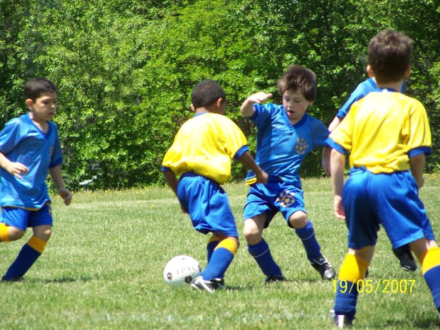 Kinder spielen Fußball auf einer Wiese mit Bäumen im Hintergrund und einem Datumsstempel in der rechten unteren Ecke.