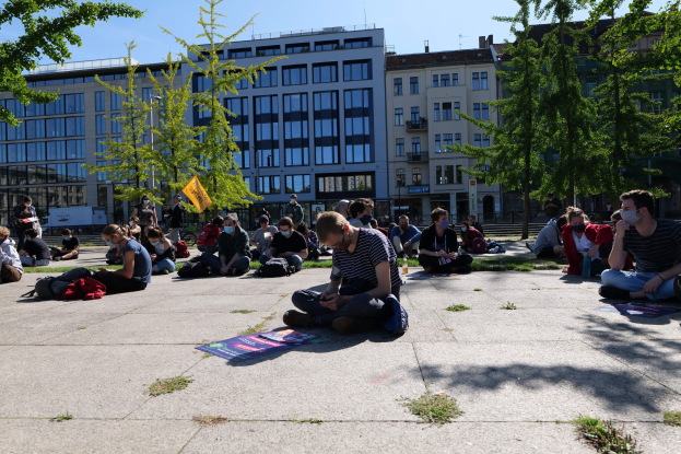Eine Gruppe von Menschen sitzt vor einem Gebäude auf dem Boden während einer Demonstration in Berlin, umgeben von Bäumen unter einem klaren blauen Himmel, wobei einige Masken tragen und im Hintergrund verstreute Gegenstände zu sehen sind.