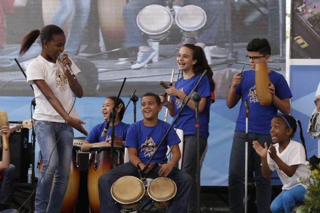 Eine Gruppe von Kindern, die auf einer Bühne Musikinstrumente spielen, mit einer Frau, die ein Mikrofon hält, in der Mitte, umgeben von Mikrofonständern und einem Banner im Hintergrund, mit Pflanzen auf der rechten Seite.