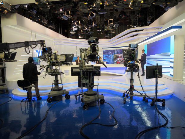 Ein Fernsehstudio mit mehreren Kameras auf dem Boden, einer sitzenden Person im Vordergrund und einigen stehenden Personen im Hintergrund, mit Deckenleuchten für eine Livesendung.