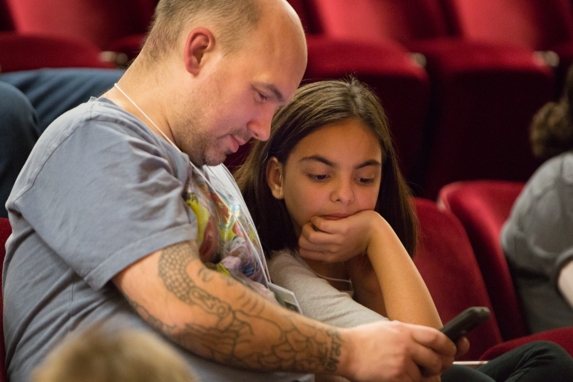 Zwei Personen, ein erwachsener Mann und ein junges Mädchen, sitzen in einem Kino und sind beide auf ein Handy konzentriert, das der Mann hält.