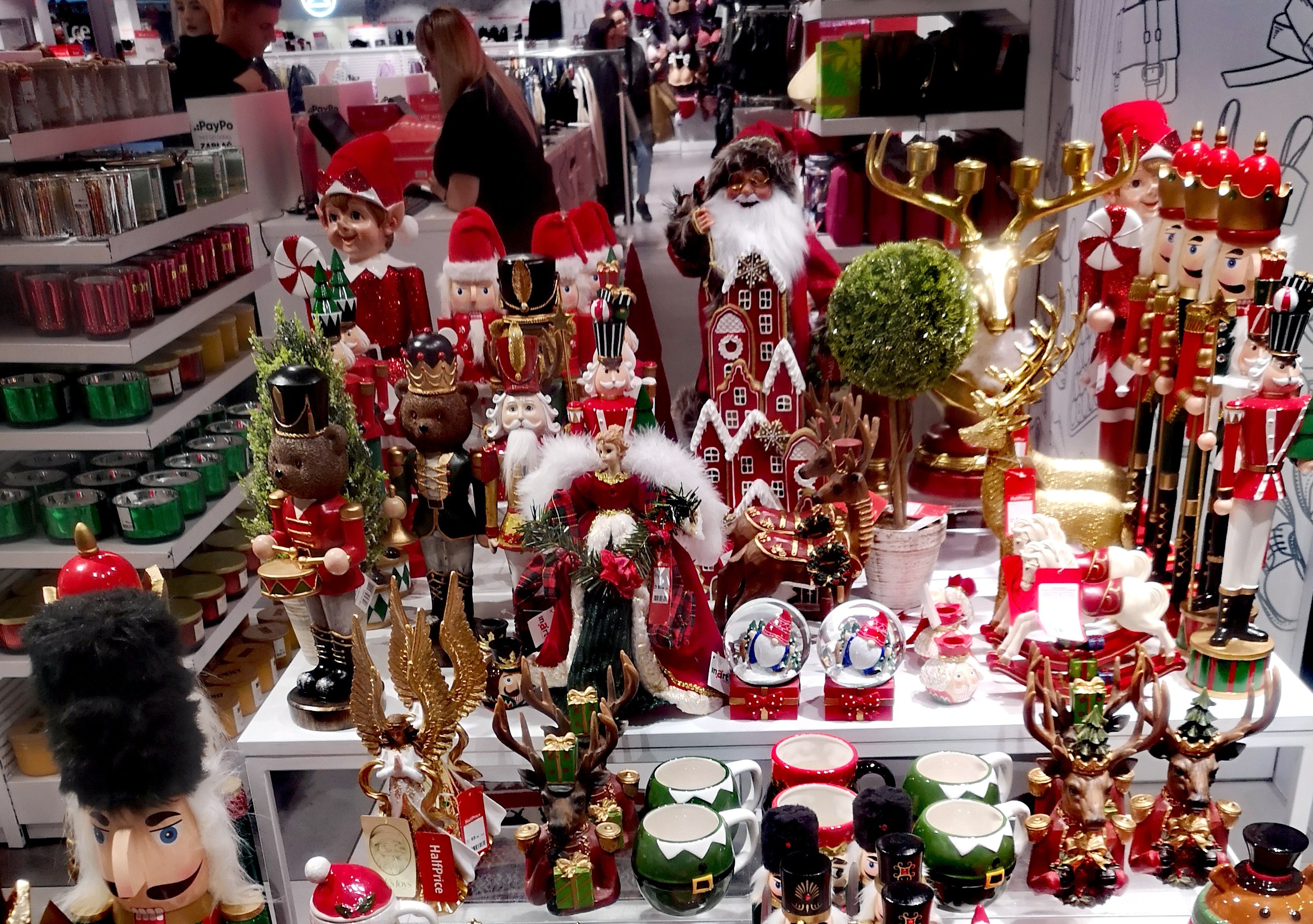 Weihnachtsdekorationen-Ausstellung mit Spielzeug, Tassen und anderen Gegenständen auf Tischen und Regalen, mit Menschen und festlichen Lichtern im Hintergrund.