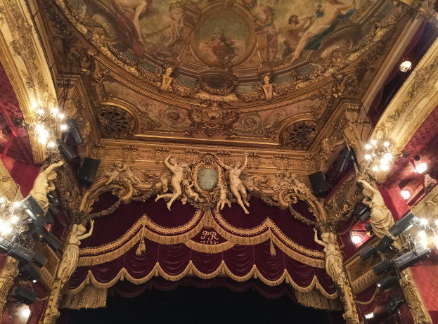 Theaterinnenraum mit rotem Vorhang, Kronleuchtern, Skulpturen, Lichtern und einer Deckenmalerei, mit einer Bühne und ein paar Menschen davor.