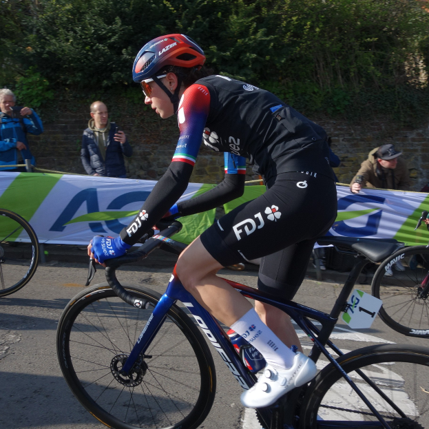 Professionelle Radrennfahrerin Katie Archibald mit Helm, Brille, Handschuhen und Schuhen fährt auf einem Fahrrad eine Straße entlang an einer Menge vorbei, mit einem Banner, einer Wand, Bäumen und einem Gebäude im Hintergrund.
