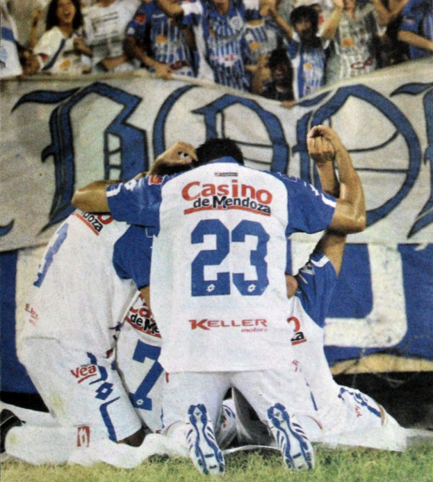 Gruppe von Füßballspielern, die vor jubelnden Fans mit einem Banner im Hintergrund auf dem Boden feiern.