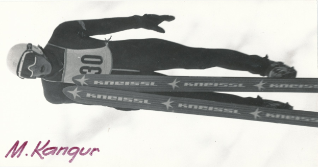 Schwarzes und weißes Foto eines Skifahrers in der Luft, der einen Helm, eine Brille, Handschuhe, Schuhe trägt und Ski hält, mit Text unten.