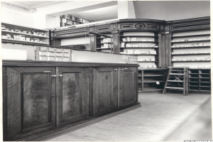 Ein Schwarz-Wei├č-Foto einer Apotheken-Inneneinrichtung mit einer Theke, Sch├Ąben, einer Leiter und Regalen voller verschiedener Gegenst├Ąnde, mit Text ├╝ber dem Bild.