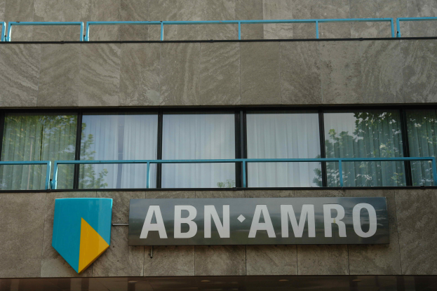 Ein Gebäude mit einer Tafel, auf der "ABN AMRO" steht, mit einer Tafel, die es als Hauptsitz angibt, einem Geländer und Glasfenstern mit Vorhängen, mit Text am unteren Rand, der eine Strafe von 1,5 Milliarden Dollar durch die Europäische Union erwähnt.