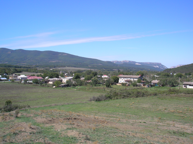 Ein kleines Dorf liegt inmitten einer grünen Wiese umgeben von Bäumen und Pflanzen, mit majestätischen Bergen und Wolken im Hintergrund.